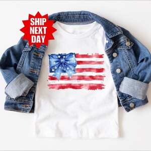 Può includere: T-shirt bianco con un disegno ad acquerello della bandiera americana e un fiocco blu. La bandiera ha strisce rosse e bianche e un fiocco blu con stelle bianche. Una giacca di jeans è drappeggiata sulla maglietta.