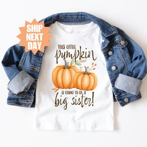以下が含まれることがあります： 「This little pumpkin is going to be a big sister!」の文字とカボチャのイラストが描かれた白いTシャツ。デニムジャケットがTシャツの上に重ねられています。