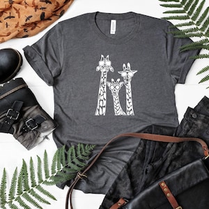 May include: Charcoal grey t-shirt with a white illustration of three giraffes wearing sunglasses. The shirt is shown with black boots, a leather bag, and dark jeans. A fern and a patterned scarf are also in the image.