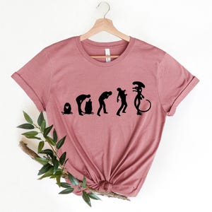 May include: A pink t-shirt with a black graphic of the evolution of the alien from the movie Alien. The graphic shows a series of silhouettes, starting with an egg and ending with the alien.