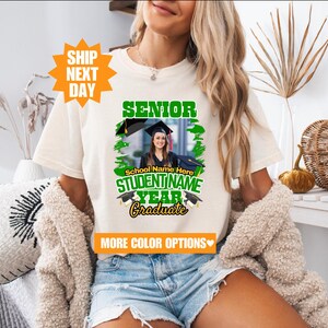 May include: White t-shirt with a green and black graphic that says "Senior" and "Graduate". The graphic also includes a photo of a person wearing a graduation cap and gown. The text "School Name Here" and "STUDENT NAME" are also included in the graphic.