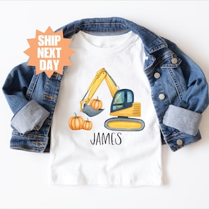 Pode incluir: Camiseta branca com um desenho de escavadeira amarela, abóboras e o nome "JAMES". A camiseta é exibida em uma jaqueta jeans. O texto "SHIP NEXT DAY" está em uma estrela laranja.