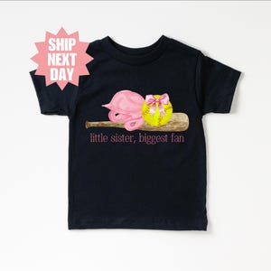 May include: Black t-shirt with a pink softball helmet, a yellow softball with a pink bow, and a brown baseball bat. The text "little sister, biggest fan" is printed in pink on the shirt.