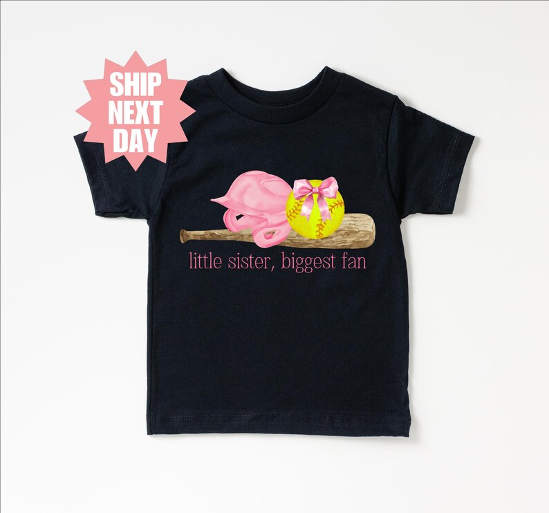 May include: Black t-shirt with a pink softball helmet, a yellow softball with a pink bow, and a brown baseball bat. The text "little sister, biggest fan" is printed in pink on the shirt.