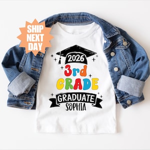 Op de afbeelding: Wit T-shirt met een kleurrijk "3rd Grade Graduate" ontwerp, met een afstudeerhoed en het jaar 2026. Het shirt wordt gecombineerd met een blauw spijkerjasje. De naam "Sophia" staat op het shirt.