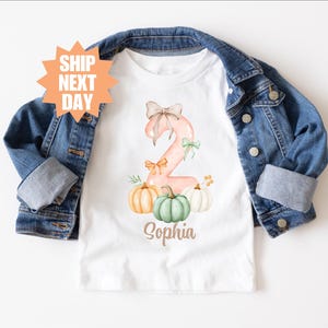 May include: A white short-sleeved t-shirt featuring a large pink number two decorated with bows and three pumpkins in shades of orange, green, and white. The name Sophia is printed below in brown script. A blue denim jacket is layered over the shirt.
