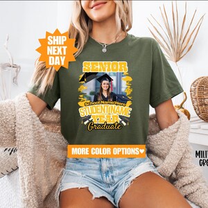 May include: A green t-shirt with a yellow graphic that says "Senior" and has a photo of a graduate in a cap and gown. The graphic also includes the text "School Name Here", "STUDENT NAME", "YEAR", and "Graduate".