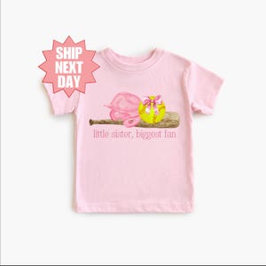 May include: Pink t-shirt with a softball, bat, and catcher's mitt graphic. The text "little sister, biggest fan" is printed below the graphic.