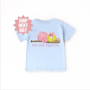 May include: Light blue t-shirt with a pink softball helmet, a yellow softball with a pink bow, and a brown baseball bat. The text "little sister, biggest fan" is printed on the shirt.