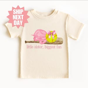 May include: A white t-shirt with a pink softball helmet, a yellow softball with a pink bow, and a brown baseball bat. The text "little sister, biggest fan" is printed on the shirt.