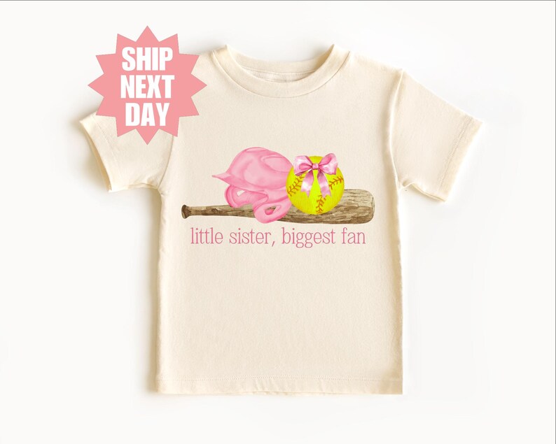 May include: A white t-shirt with a pink softball helmet, a yellow softball with a pink bow, and a brown baseball bat. The text "little sister, biggest fan" is printed on the shirt.