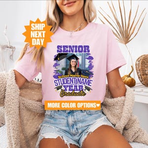 May include: Pink t-shirt with a purple and gold graphic that says "Senior" and "Graduate". The graphic also includes a photo of a person wearing a graduation cap and gown. The text "School Name Here" and "STUDENTNAME YEAR" is also included in the graphic.