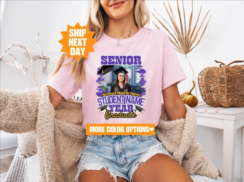 May include: Pink t-shirt with a purple and gold graphic that says "Senior" and "Graduate". The graphic also includes a photo of a person wearing a graduation cap and gown. The text "School Name Here" and "STUDENTNAME YEAR" is also included in the graphic.