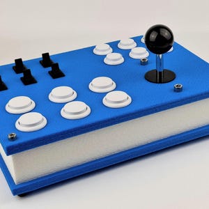 May include: A blue and white arcade game controller with a black joystick and white buttons. The controller has a rectangular shape with a blue top and white sides. Black switches are also visible.