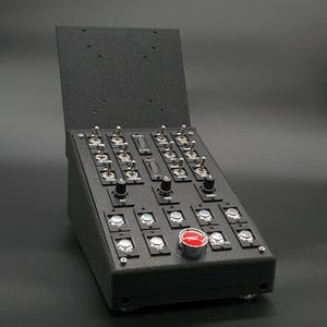 May include: Black control panel with numerous toggle switches, knobs, and push buttons. The panel has a hinged top and a prominent red button. The switches are labeled with various functions.