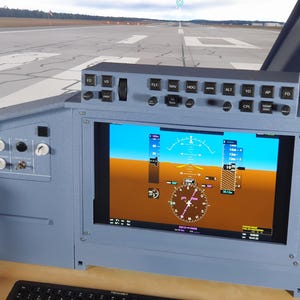 Op de afbeelding: Een lichtblauw bedieningspaneel van een vluchtsimulator met een groot scherm waarop vlieginstrumenten worden weergegeven. Het paneel heeft verschillende knoppen en schakelaars en het scherm toont een gedetailleerd beeld van de houding en navigatiegegevens van een vliegtuig. De achtergrond toont een luchthavenbaan.