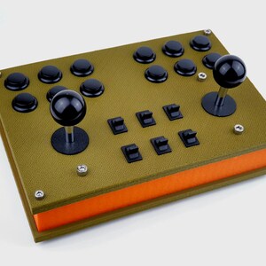 Puede incluir: Un mando de juego de estilo arcade verde y naranja con dos joysticks y varios botones. El mando tiene una superficie texturizada y está hecho de madera.