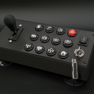 Pode incluir: Uma caixa de controlo retangular preta com um joystick à esquerda, uma grelha de dez botões de pressão circulares com ícones brancos, um botão de perigo triangular vermelho, um interruptor basculante e uma chave de ignição com porta-chaves.