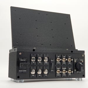May include: Black control panel with multiple switches, buttons, and labels, including "GEAR UP," "GEAR DOWN," and "IGNITION." The panel has a hinged top and is mounted on silver feet. The panel is designed for flight simulation.
