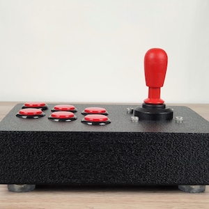 Könnte beinhalten: Ein schwarzer Arcade-Gamecontroller mit einem roten Joystick und sechs roten Knöpfen. Der Controller hat eine strukturierte Oberfläche und ist auf vier kleinen, grauen Füßen montiert. Der Joystick befindet sich mittig auf der rechten Seite.