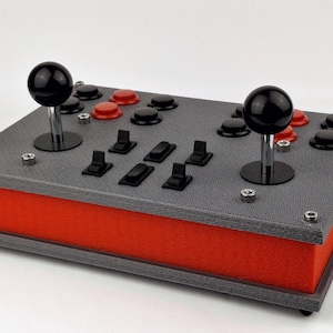 Puede incluir: Un mando de arcade gris y rojo con dos joysticks y varios botones. El mando tiene una superficie texturizada y una base roja.