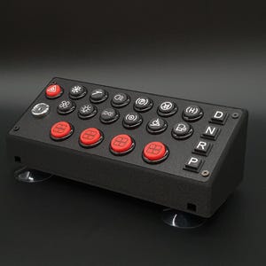 May include: A black rectangular control panel featuring a variety of circular and square buttons. The top rows contain black buttons with white icons, while the bottom row features four red square buttons and four black square buttons labeled D, N, R, P.