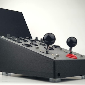 Puede incluir: Panel de control de arcade negro con dos joysticks, varios botones y un botón rojo. El panel tiene una cubierta negra que se puede levantar.