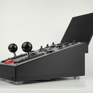 Op de afbeelding: Zwart bedieningspaneel met twee zwarte joysticks en een rode knop. Het paneel heeft een getextureerd oppervlak en een zwarte kap die gedeeltelijk open is.