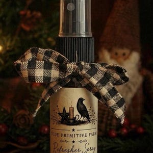 May include: A clear plastic spray bottle with a black pump top and a black and tan buffalo check ribbon tied around the neck. The label features a black crow, a star, and a candle illustration with the text Olde Primitive Farm Refresher Spray Country Store.