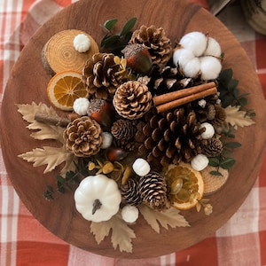 May include: A circular wooden tray centerpiece featuring an arrangement of pinecones, acorns, dried orange slices, cinnamon sticks, white cotton bolls, small white pumpkins, and beige faux leaves, creating a textured, multi-layered seasonal display.