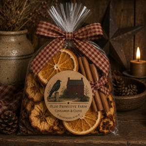 May include: A clear plastic bag filled with dried orange slices, cinnamon sticks, and star anise. A brown circular label features a black crow, a red house, and the text OLDE PRIMITIVE FARM CINNAMON & CLOVE HANDMADE IN RI. A red and tan gingham ribbon is tied at the top.
