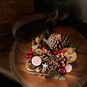 May include: A wooden tray filled with festive Christmas decorations, including pine cones, cinnamon sticks, red berries, and gingerbread cookies. The tray is adorned with a red and white striped candy cane and a red and white striped candle.