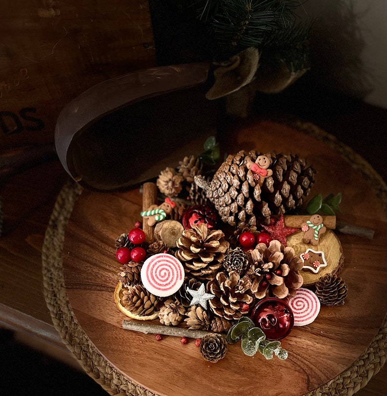 May include: A wooden tray filled with festive Christmas decorations, including pine cones, cinnamon sticks, red berries, and gingerbread cookies. The tray is adorned with a red and white striped candy cane and a red and white striped candle.
