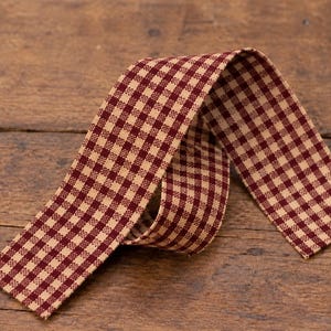 May include: A strip of fabric ribbon featuring a small-scale gingham check pattern in deep burgundy red and cream. The ribbon is folded into a loose, overlapping loop, displaying the consistent checkered design on its flat, woven surface.