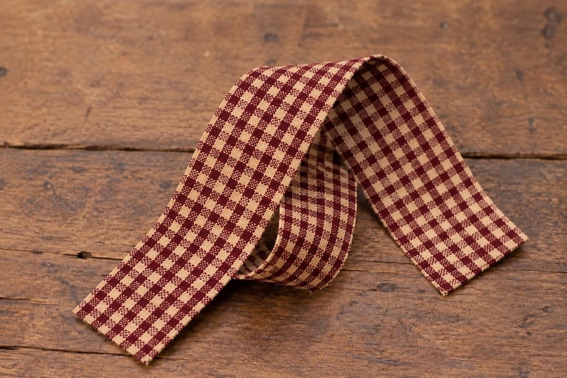 May include: A strip of fabric ribbon featuring a small-scale gingham check pattern in deep burgundy red and cream. The ribbon is folded into a loose, overlapping loop, displaying the consistent checkered design on its flat, woven surface.