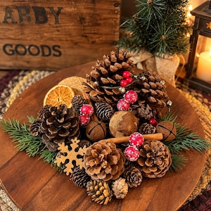 May include: A circular wooden tray centerpiece featuring an arrangement of brown pinecones, dried orange slices, cinnamon sticks, small jingle bells, red berries, and a wooden snowflake cutout, all accented with green evergreen sprigs.