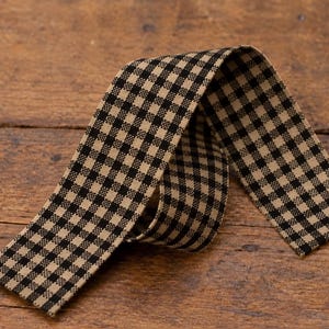May include: A length of fabric ribbon featuring a black and tan gingham check pattern. The ribbon is folded into a loose loop, displaying the woven texture of the material and the consistent grid design across the entire surface of the strip.