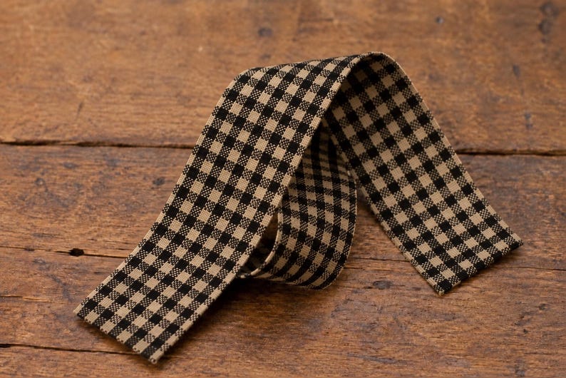 May include: A length of fabric ribbon featuring a black and tan gingham check pattern. The ribbon is folded into a loose loop, displaying the woven texture of the material and the consistent grid design across the entire surface of the strip.