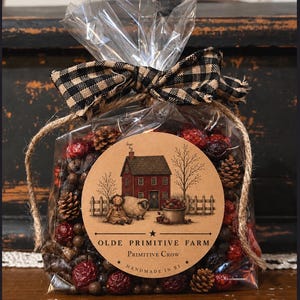 May include: A clear plastic bag filled with potpourri containing small pinecones and red berries, tied with a black and tan buffalo check ribbon. A round tan label features a red house, a sheep, and text reading OLDE PRIMITIVE FARM PRIMITIVE CROW HANDMADE IN RI.