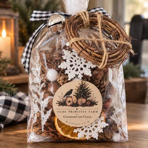 May include: A clear plastic bag filled with pinecones, white felt snowflakes, and dried orange slices. The bag is tied with a black and white gingham ribbon, a silver jingle bell, and a small grapevine wreath. A round label reads OLDE PRIMITIVE FARM CRANBERRY AND CEDAR.