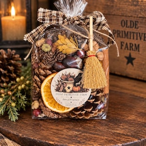 May include: A clear plastic bag filled with autumn potpourri including pinecones, dried orange slices, acorns, and small pumpkins. The bag is tied with a brown plaid ribbon and a miniature broom, featuring a label that reads OLDE PRIMITIVE FARM APPLE PUMPKIN.
