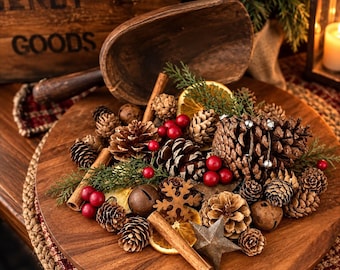 May include: A wooden scoop rests on a round wooden board filled with a collection of pinecones, cinnamon sticks, dried orange slices, red berries, metal jingle bells, a metal snowflake ornament, and a metal star, accented with green pine sprigs.