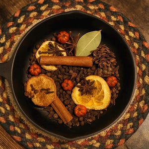 May include: A black cast iron skillet filled with dried orange slices, cinnamon sticks, star anise, cloves, bay leaf, small orange pumpkins, and a pine cone. The skillet sits on a decorative, multi-colored woven placemat.