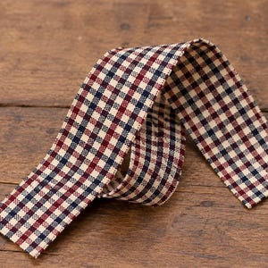 May include: A length of fabric ribbon featuring a repeating gingham check pattern in dark navy blue, deep red, and off-white. The ribbon is folded into a loose loop, displaying the woven texture of the material across its entire surface.