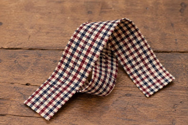 May include: A length of fabric ribbon featuring a repeating gingham check pattern in dark navy blue, deep red, and off-white. The ribbon is folded into a loose loop, displaying the woven texture of the material across its entire surface.