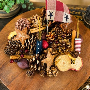 May include: A decorative arrangement featuring pinecones, cinnamon sticks, dried fruit slices, and two metal stars. It includes a small fabric flag with a red and white checkered pattern and blue stars, alongside cylindrical ornaments with patriotic motifs.