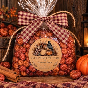 May include: A clear plastic bag filled with small orange pumpkin-shaped items, tied with a red and tan gingham ribbon. A circular label features a black crow, a basket, and the text OLDE PRIMITIVE FARM PUMPKIN SPICE HANDMADE IN RHODE ISLAND.