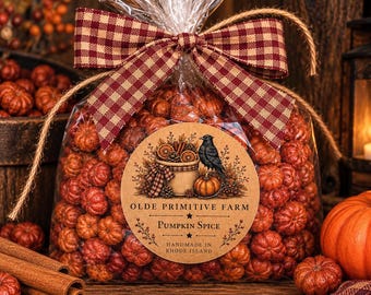May include: A clear plastic bag filled with small orange pumpkin-shaped items, tied with a red and tan gingham ribbon. A circular label features a black crow, a basket, and the text OLDE PRIMITIVE FARM PUMPKIN SPICE HANDMADE IN RHODE ISLAND.