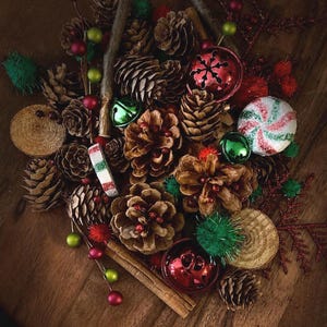 May include: A holiday arrangement featuring brown pinecones, red and green jingle bells, cinnamon sticks, wooden slices, and a peppermint candy ornament. The collection includes small red and green pom-poms and glittery accents.
