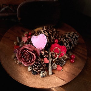 May include: A wooden plate displays a decorative arrangement of pine cones, dried rose, and felt hearts. The arrangement includes a large, dried burgundy rose, small red berries, and a pink felt heart.
